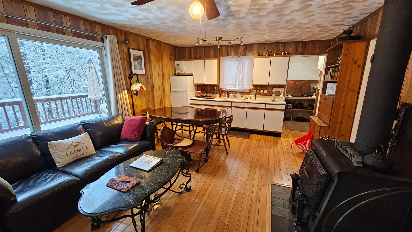 Open living room with leather sofa, wood stove, and kitchen, snowy woods out the window